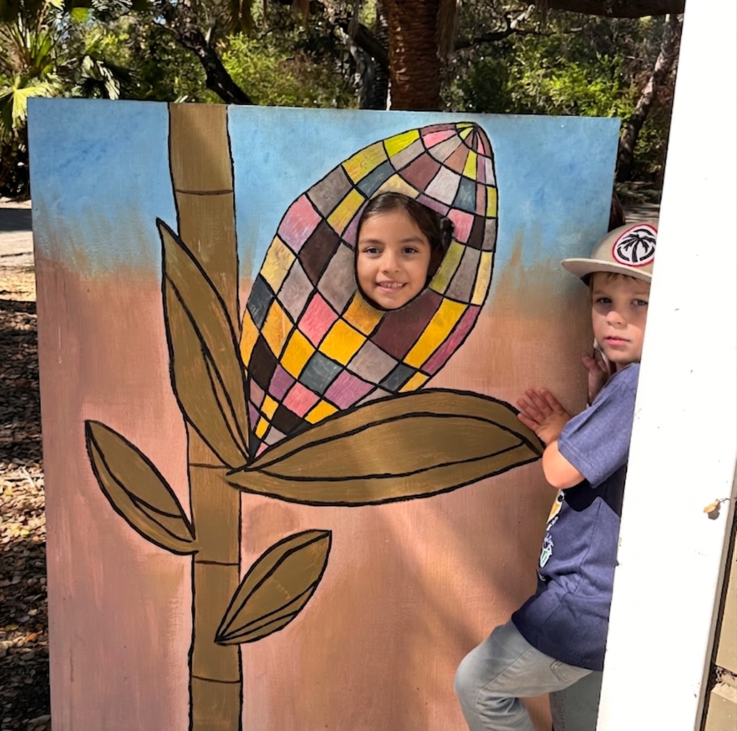 Two children peeking through a hand-painted flower photo cutout, laughing