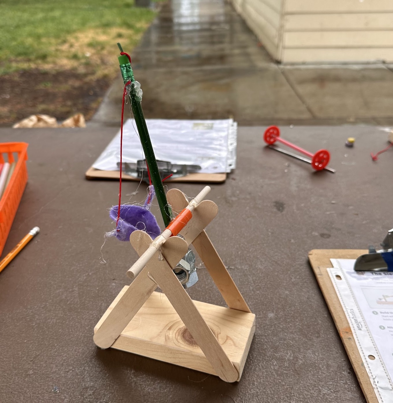 Parents and children huddled over wooden tables in the rain, collaborating to assemble complex wooden trebuchets.