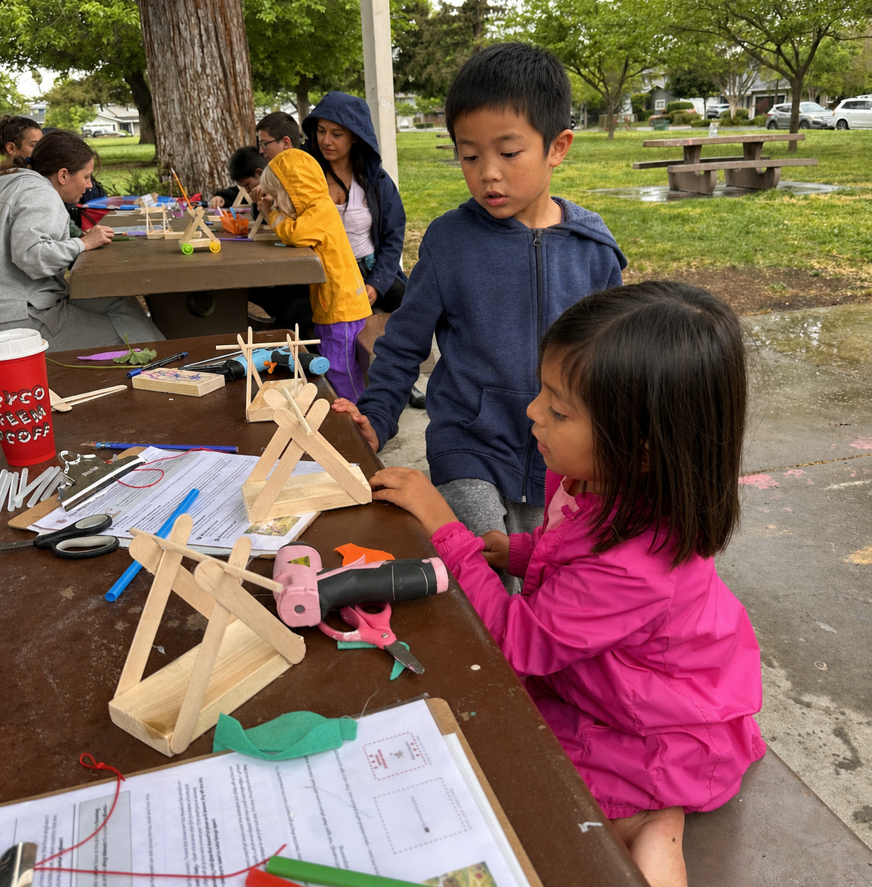A close-up of a child and parent adjusting the trigger mechanism of a wooden trebuchet.